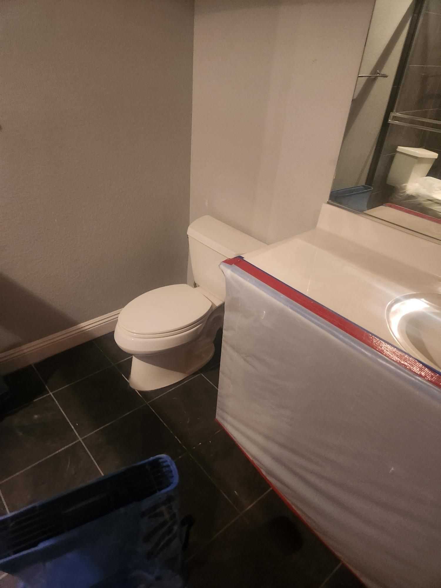 A small bathroom with a toilet, vanity, and black tile floor. The vanity is covered in a plastic sheet.