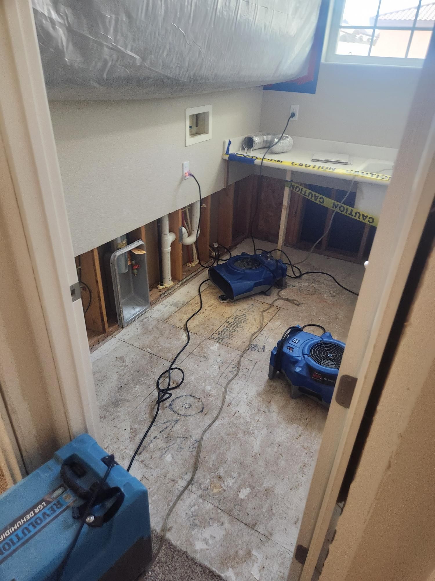 Interior view of a room undergoing water damage restoration, with exposed wall studs, drying fans, and caution tape.