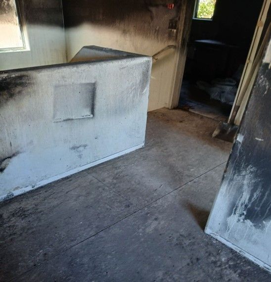 Interior of a room damaged by fire, with soot covering walls and floor. A doorway and window are visible in the background.