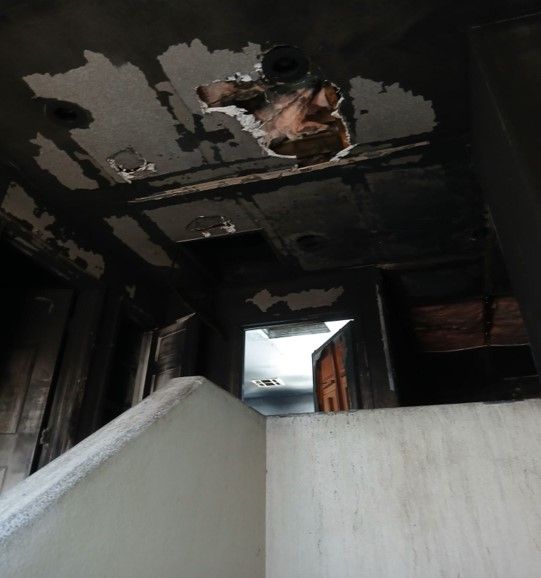 Interior of a damaged building after a fire. Smoke and debris cover the ceiling and walls; a staircase is visible.