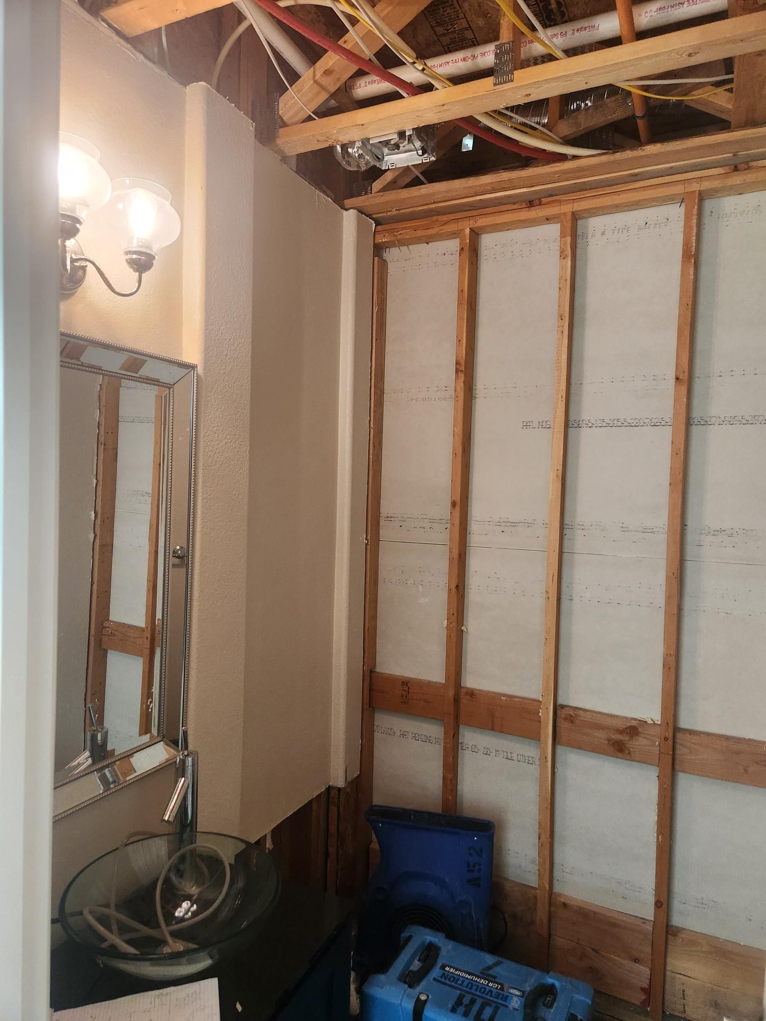 Bathroom in disrepair with exposed wall studs, a mirror, and a light fixture. A blue appliance sits at the base of the wall.
