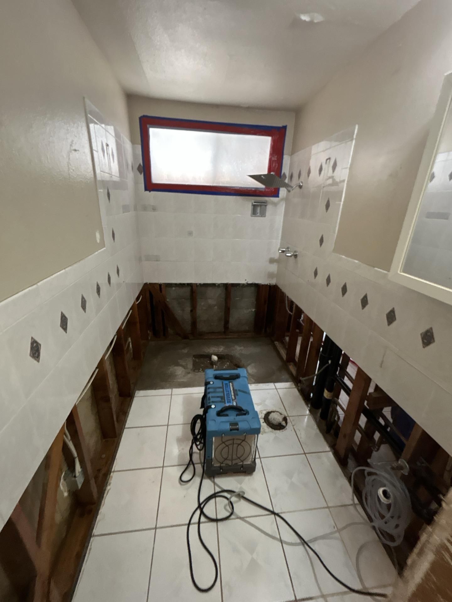 Bathroom under renovation with exposed studs, white tile, and a blue machine on the floor. The shower has a red barrier over the window.