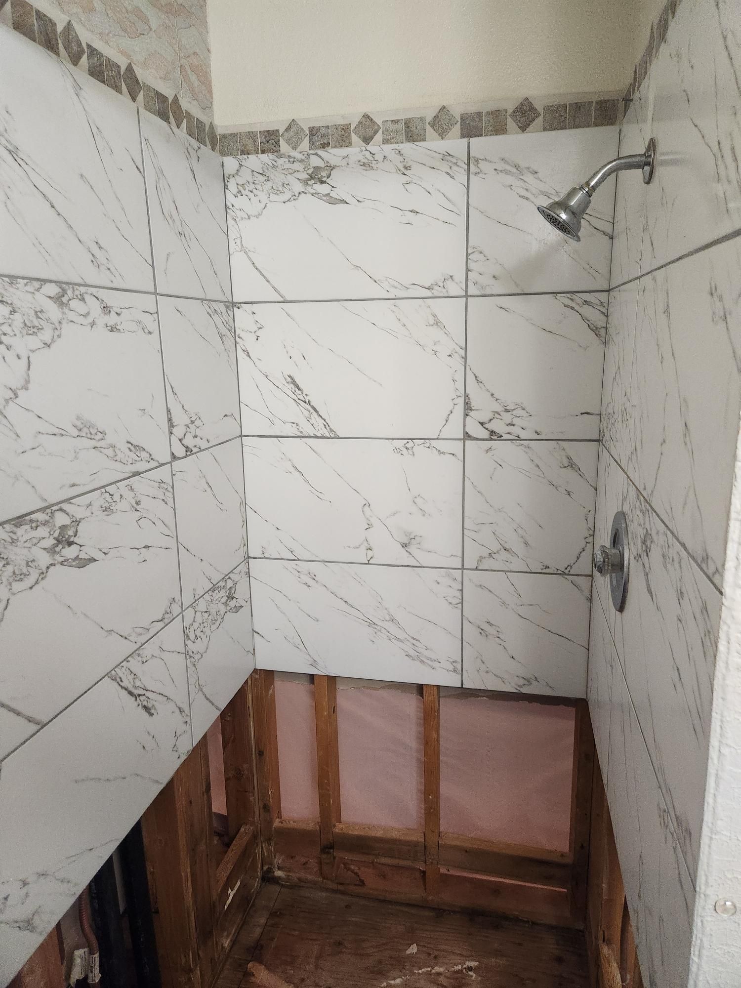 Shower stall with marble-patterned tiles, exposed wooden framing, and a silver showerhead. The lower wall is missing tiles.