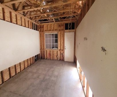 Interior view of a room under construction. Bare wooden studs are visible, with drywall partially installed on the walls. A window and door are also present.