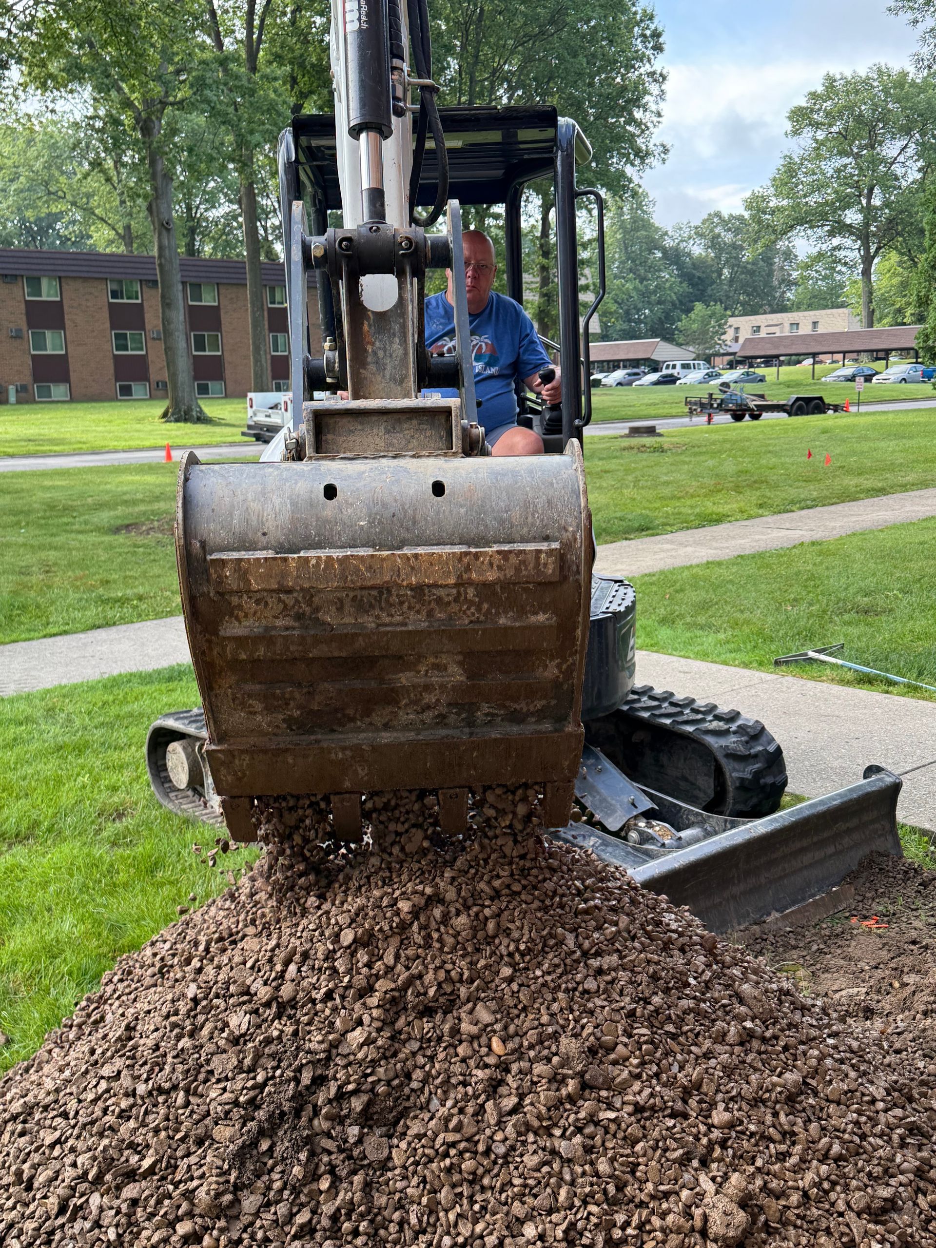 A man is driving a small excavator in front of a brick building.