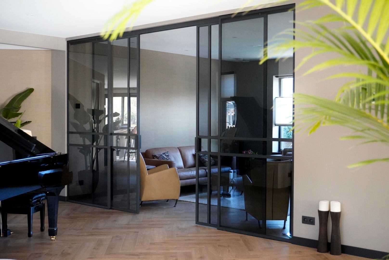Interieur met een zwarte glazen deur, die toegang geeft tot een woonkamer met een bank, een piano en planten.