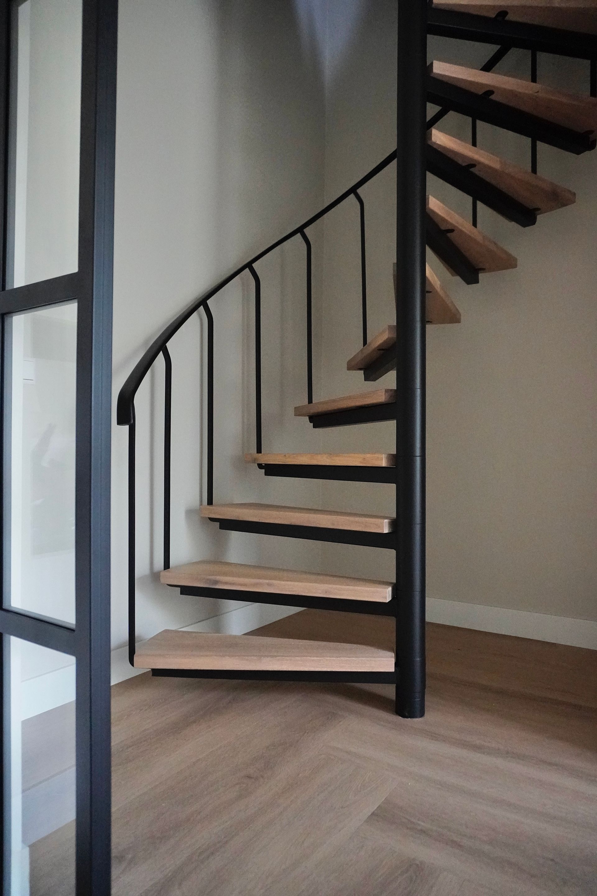 Wenteltrap met houten treden, zwarte leuning en metalen steun. De ruimte heeft een houten vloer en een lichtgekleurde muur.