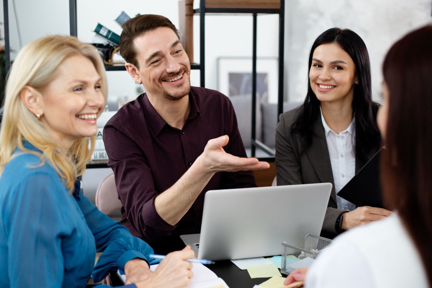 A professional team collaborates in an office, with one person gesturing while speaking to colleagues around a laptop.