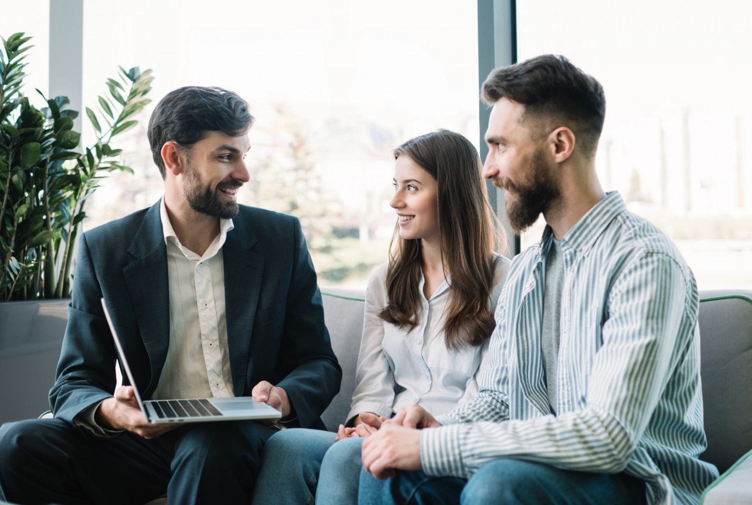 A man in a suit displays a laptop screen to a man and woman sitting together on a couch in a bright office.