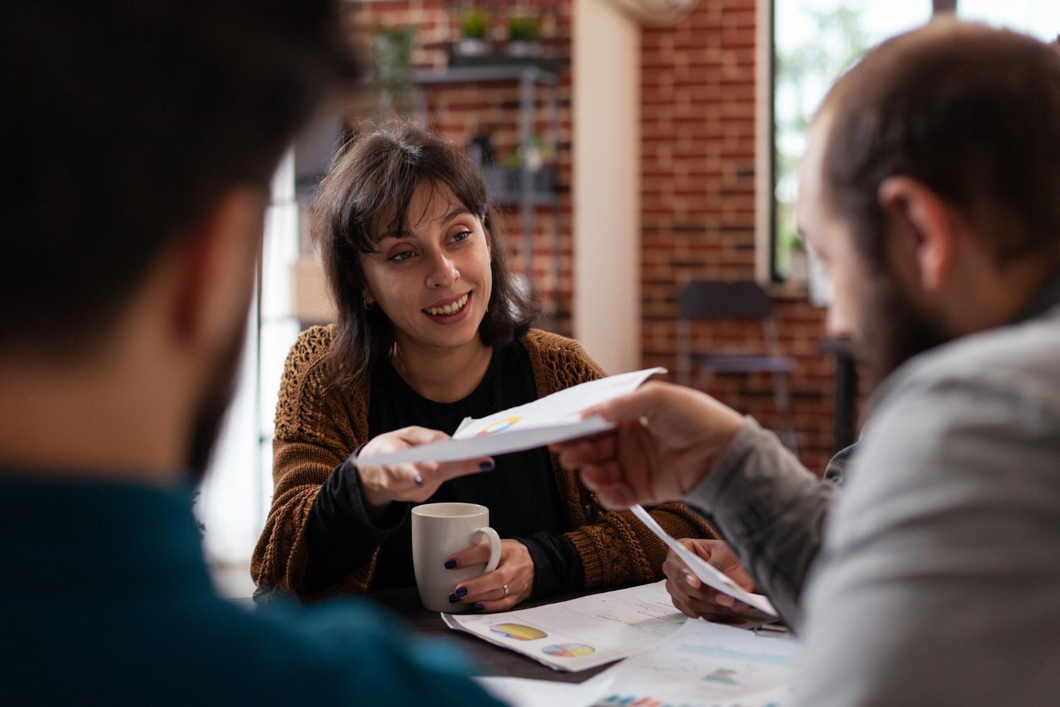 Three colleagues sit at a table in a workspace, discussing documents and charts with a coffee mug present.