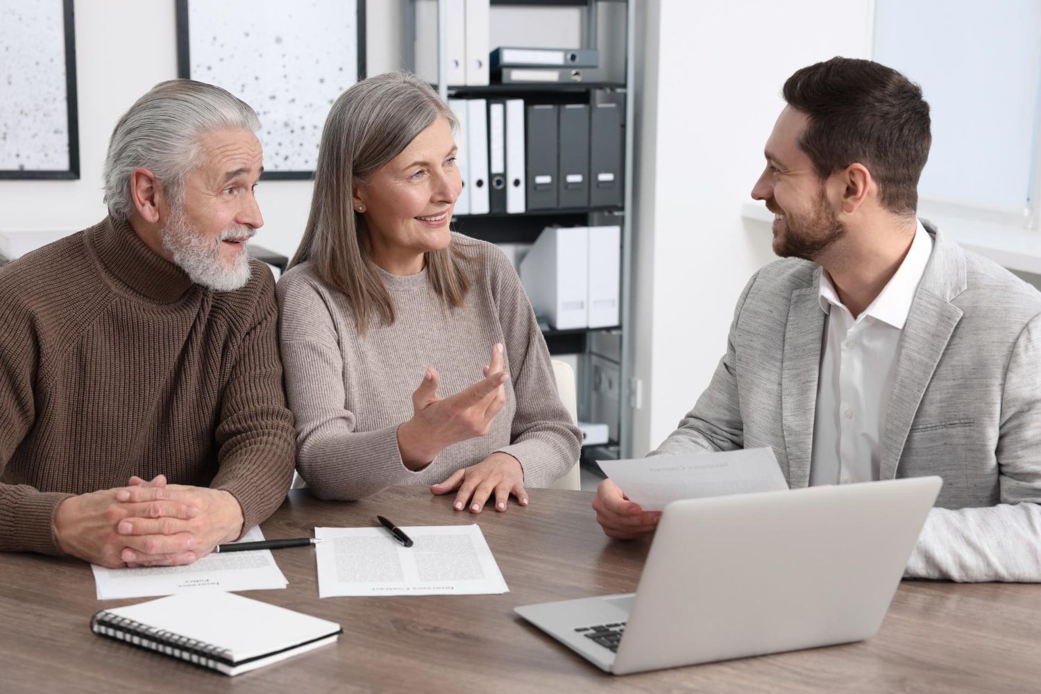 Two people sit at a desk talking to a consultant in an office with a laptop and documents.