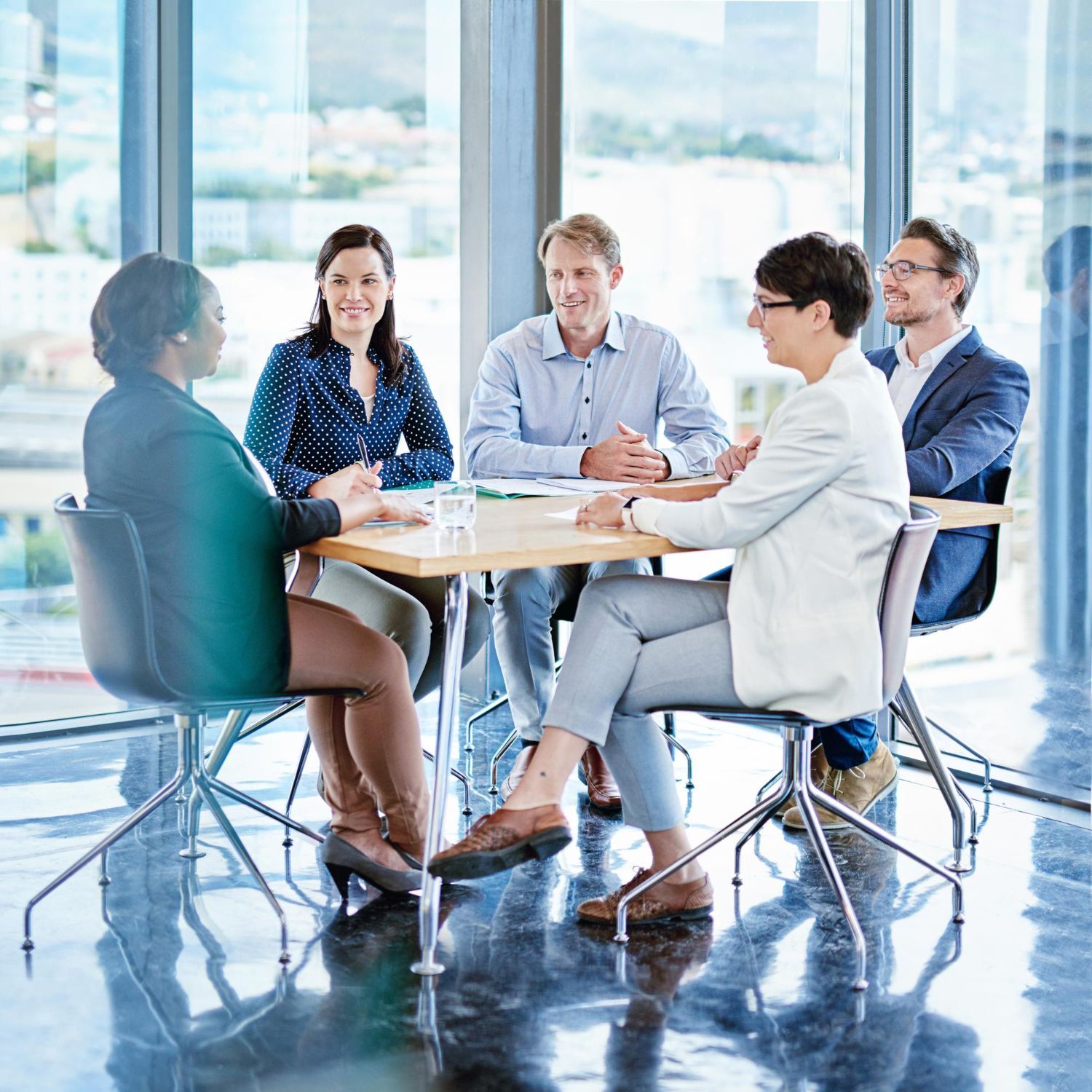 Five people sit around a wooden table in a bright, modern office conference room for a professional business meeting.