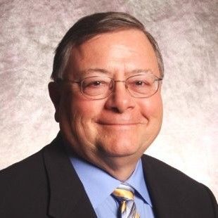 A headshot of a person wearing glasses, a dark suit, and a light blue shirt with a patterned tie against a gray background.
