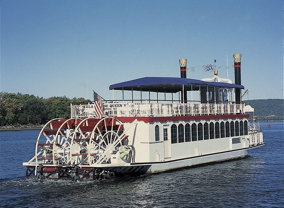 A white riverboat with a red-trimmed deck and large paddle wheel on the back, sailing on a calm river under a clear sky.