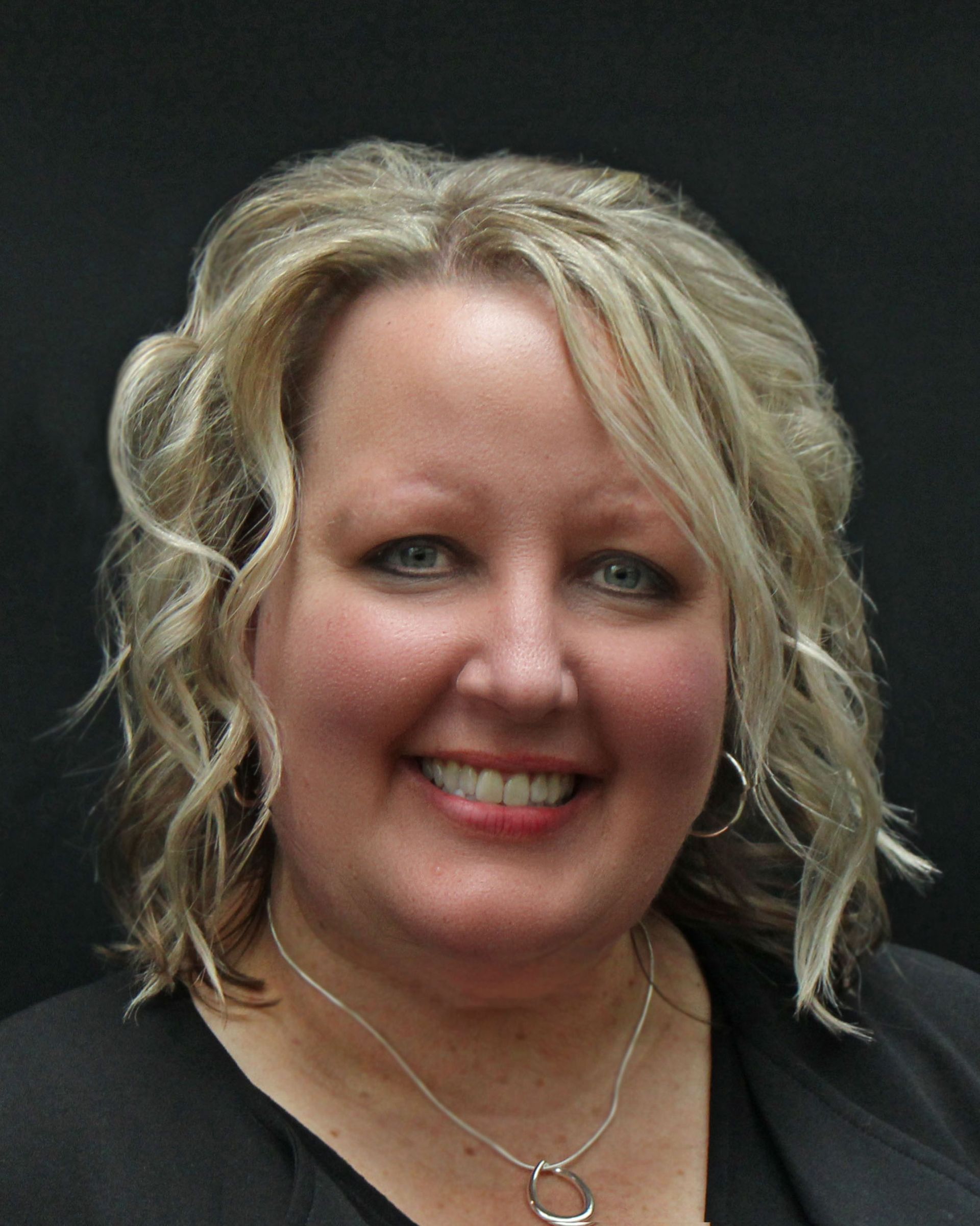 A smiling person with shoulder-length blonde wavy hair, wearing a dark top and a silver necklace against a black background.