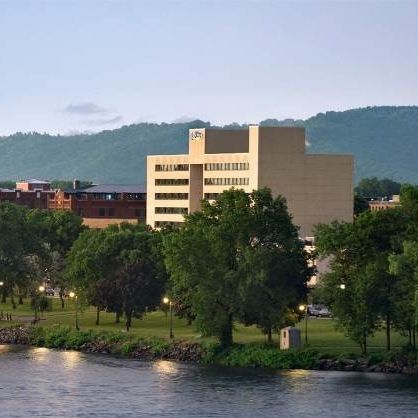 A tan mid-rise building stands behind a lush green park along a river, with mountains in the background under a blue sky.
