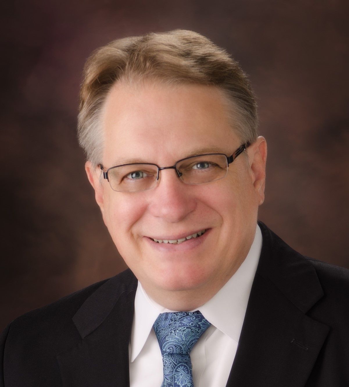 A smiling professional wearing glasses, a dark blazer, white shirt, and a blue patterned tie against a brown background.