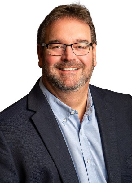 A person with short brown hair and glasses, wearing a light blue button-up shirt and a dark blazer, smiling against white.