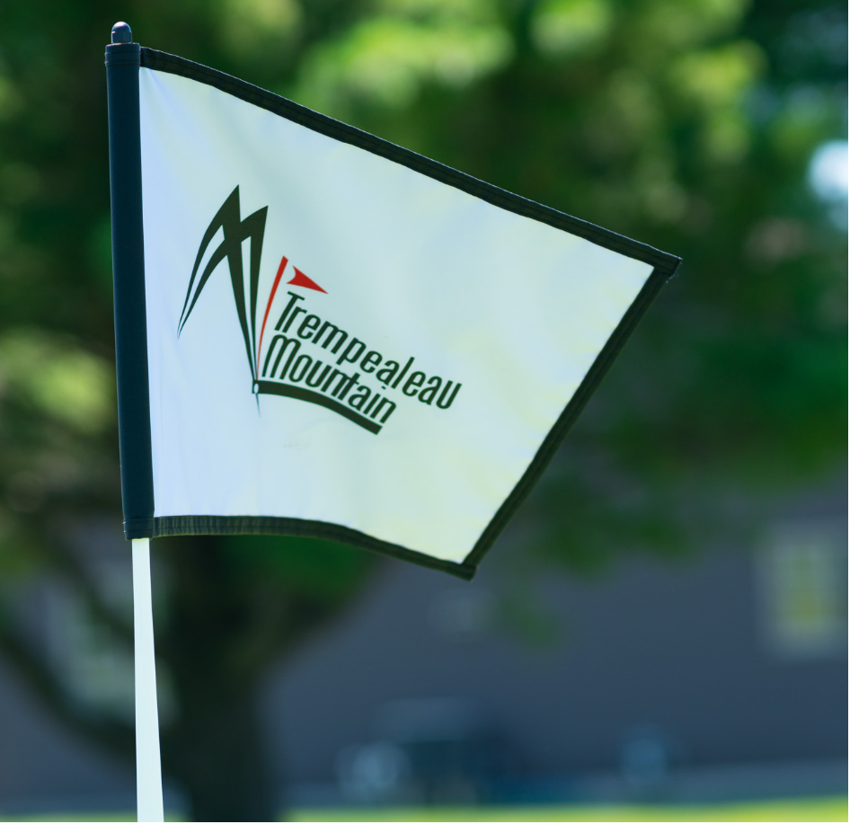 A white golf flag with the Trempealeau Mountain logo, displayed against an out-of-focus background of trees and a building.