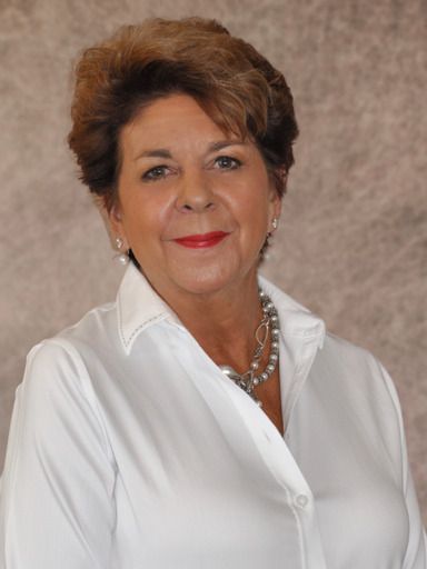 A person with short brown hair wearing a white button-down shirt and a pearl necklace, posing against a neutral backdrop.
