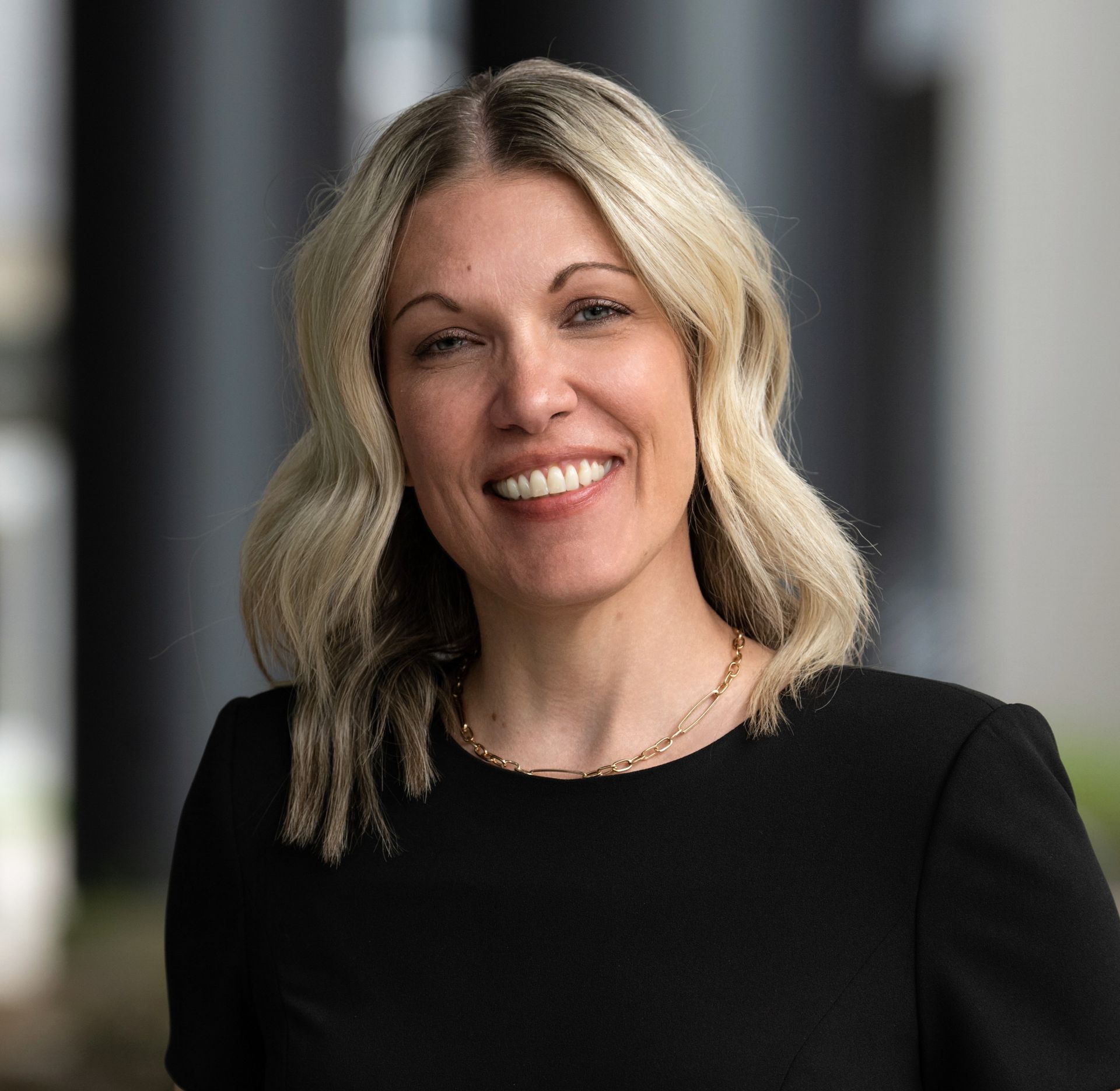 Headshot of a smiling person with shoulder-length blonde hair, wearing a black top and gold chain necklace.
