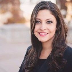 A professional headshot of a smiling person with shoulder-length wavy dark hair, wearing a dark blazer in an outdoor setting.