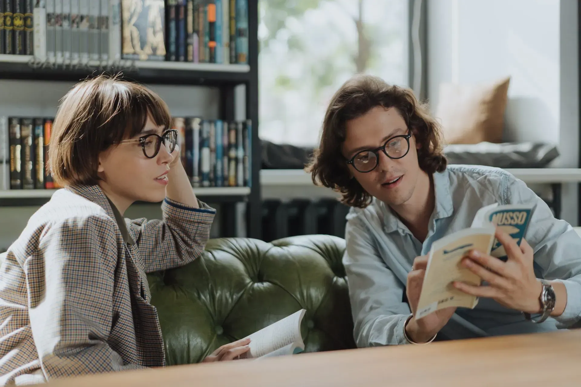 Two people sit on a green leather sofa in a library, looking at an open book together.