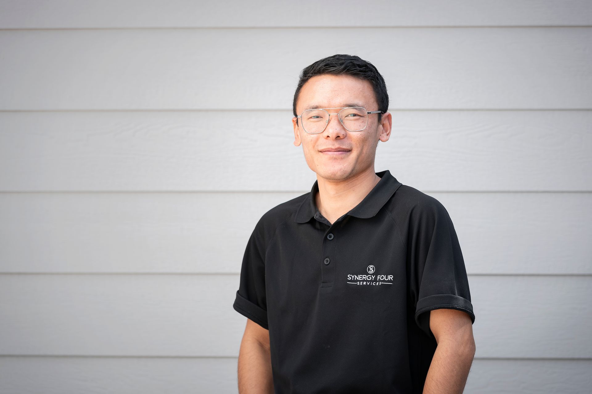 A man wearing glasses and a black polo shirt is standing in front of a white wall.