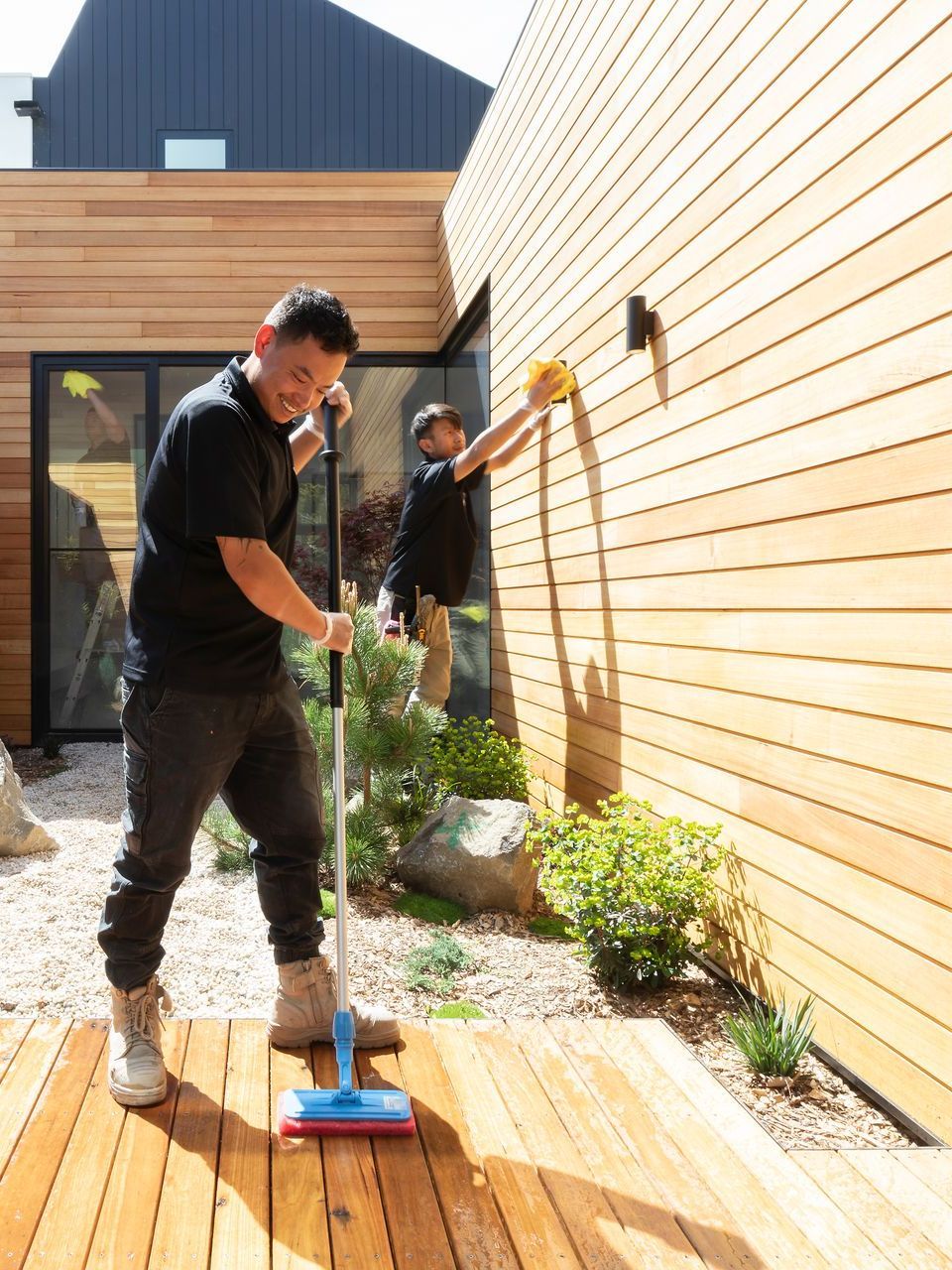 Two men are cleaning a wooden deck outside of a house.