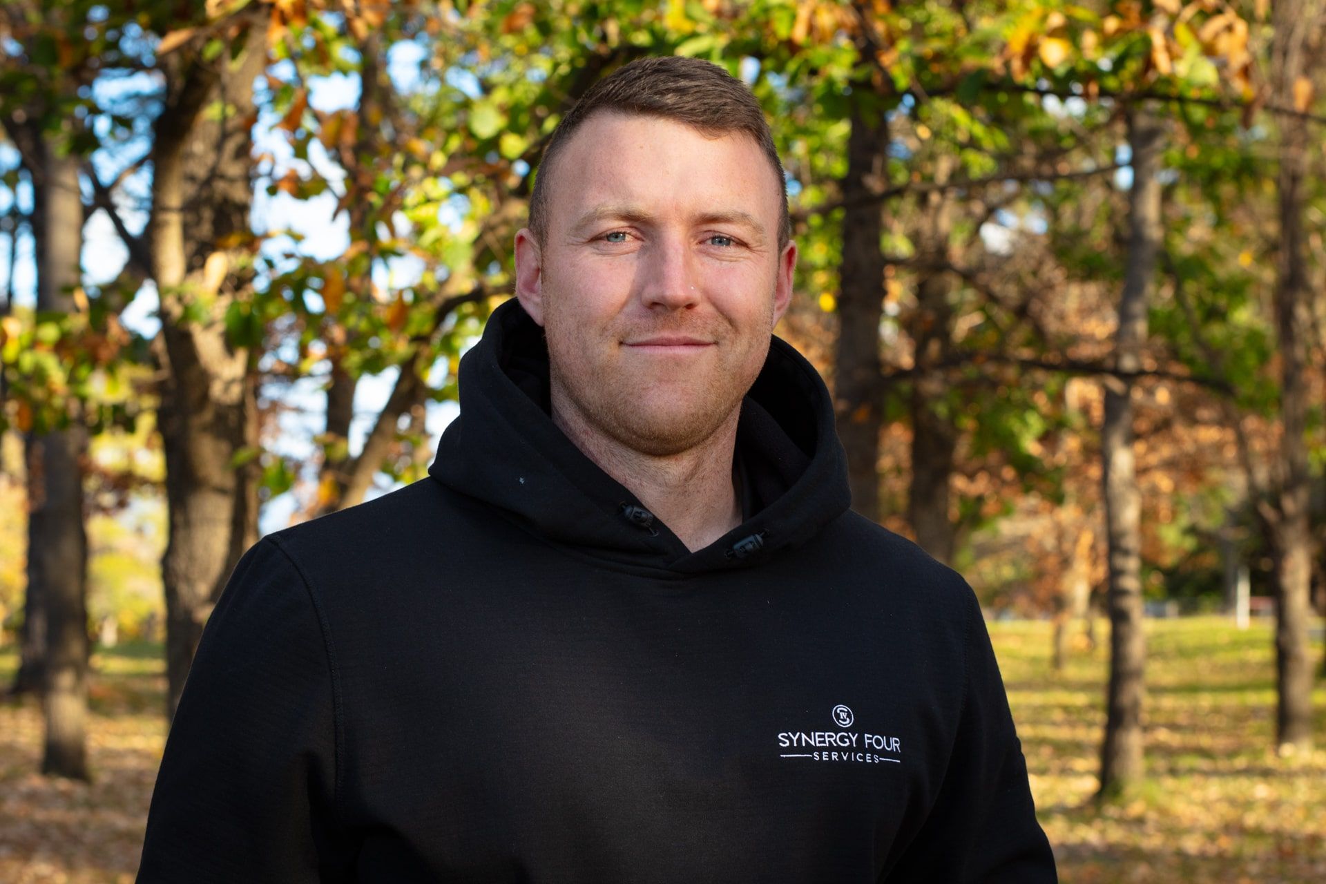 A man in a black hoodie is standing in front of a forest.