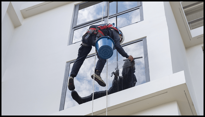Professional window cleaner suspended by rope, using safety gear while cleaning exterior windows of a high-rise building