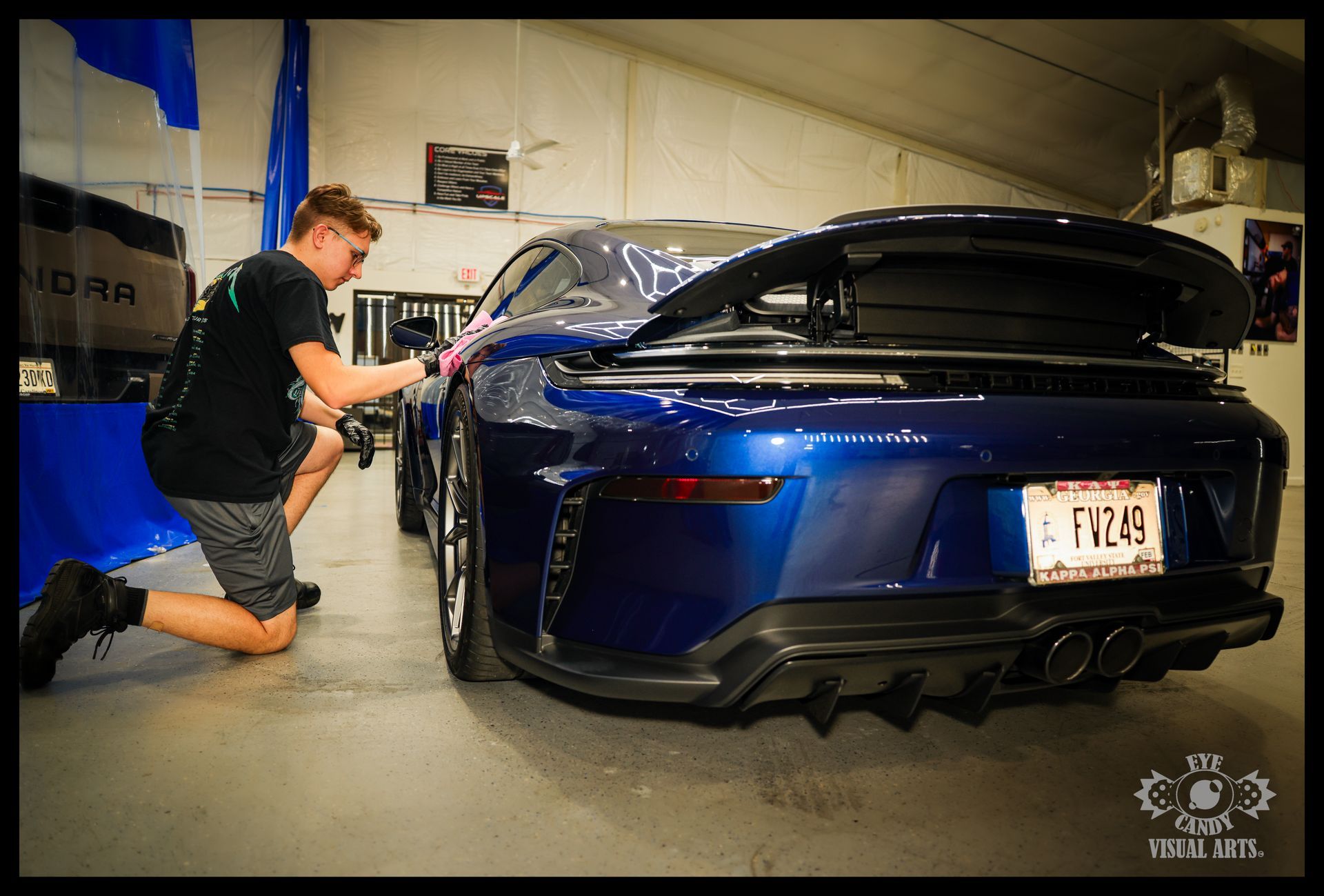 Person kneels beside a blue sports car in a garage, applying work near the rear quarter panel.