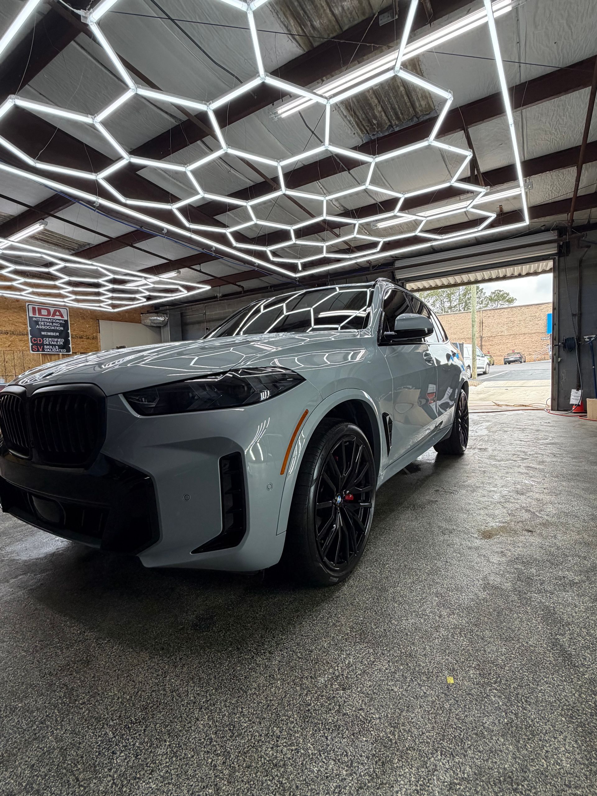 Gray BMW SUV parked under hexagon-shaped lights in a car detailing shop, open door in background.