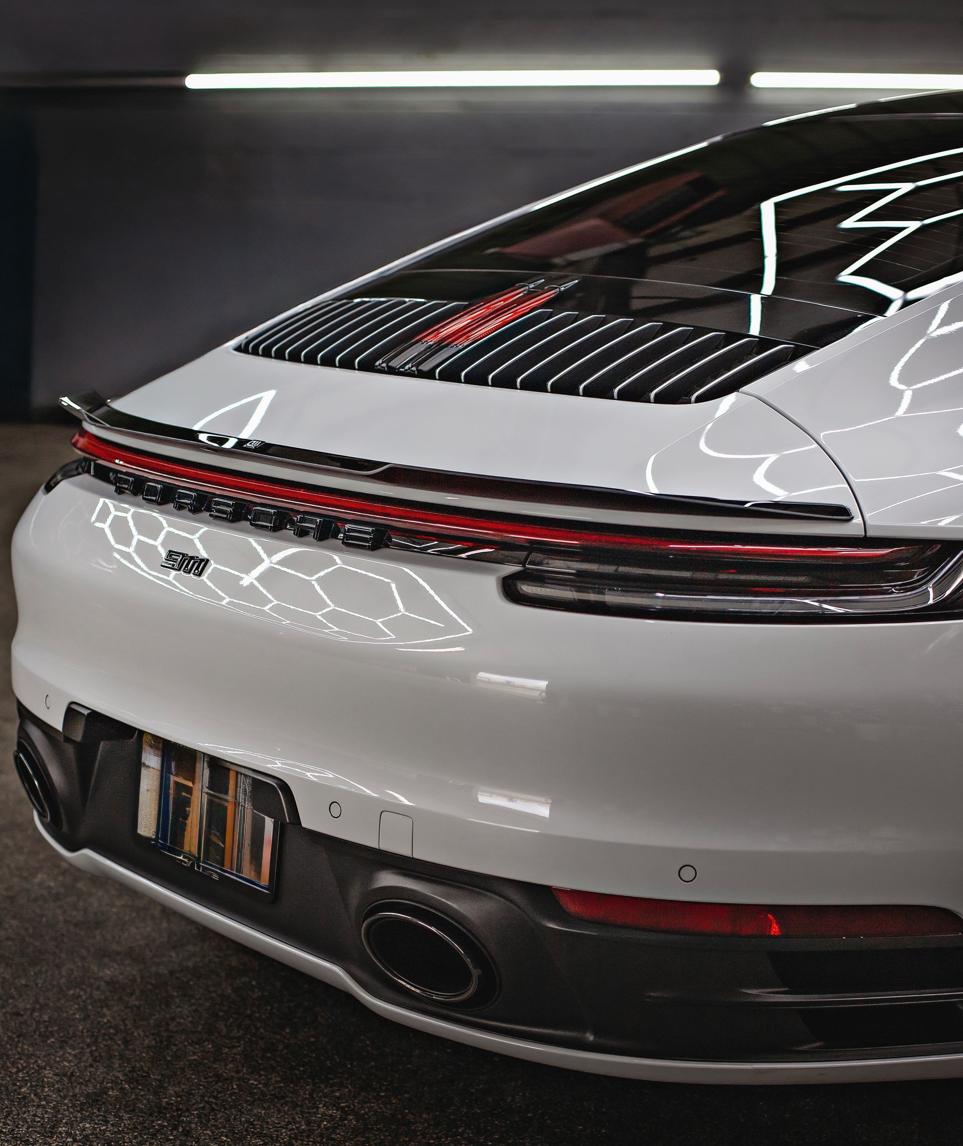 White Porsche 911 rear view, featuring black spoiler, taillights, and exhaust in a dimly lit garage.