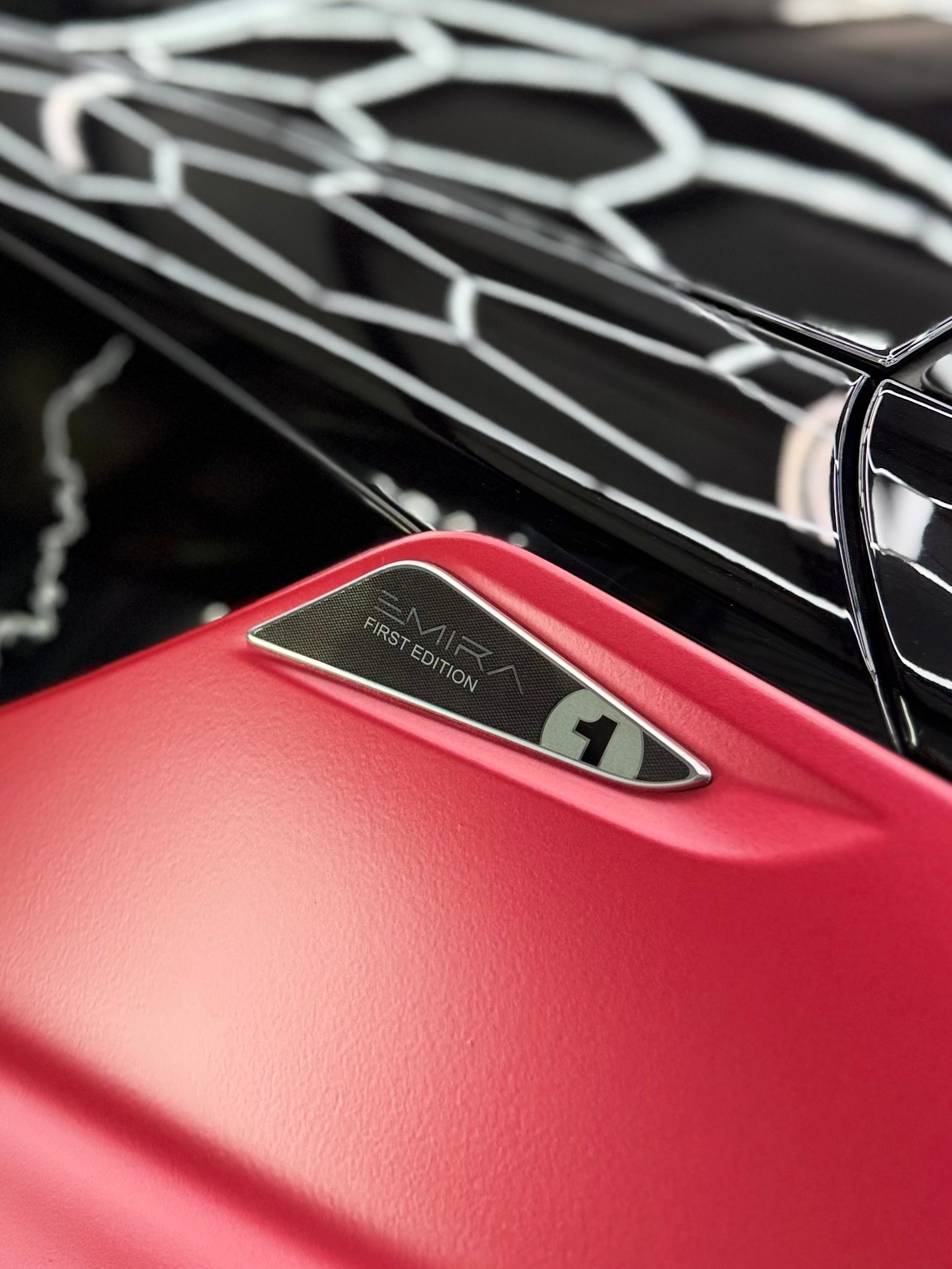 Close-up of a red sports car hood with a black honeycomb pattern on the car behind it; a small emblem.