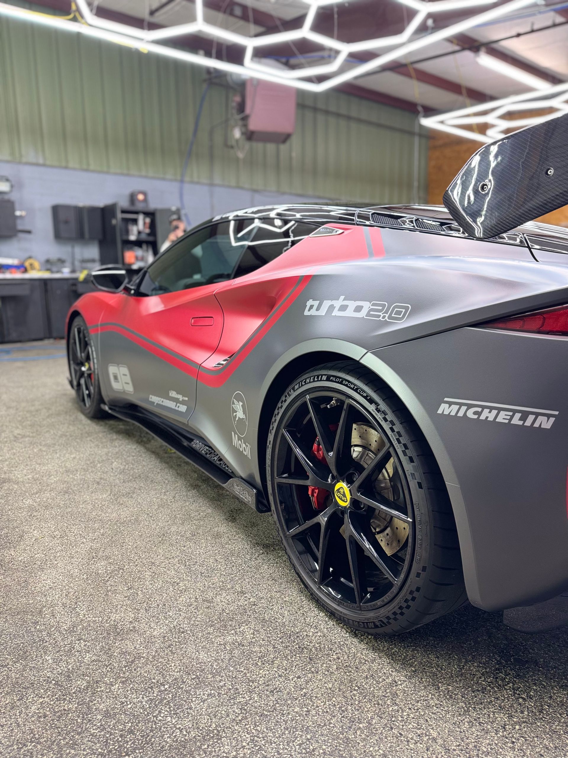 Gray and red sports car with black wheels in a garage, under hexagonal lights.
