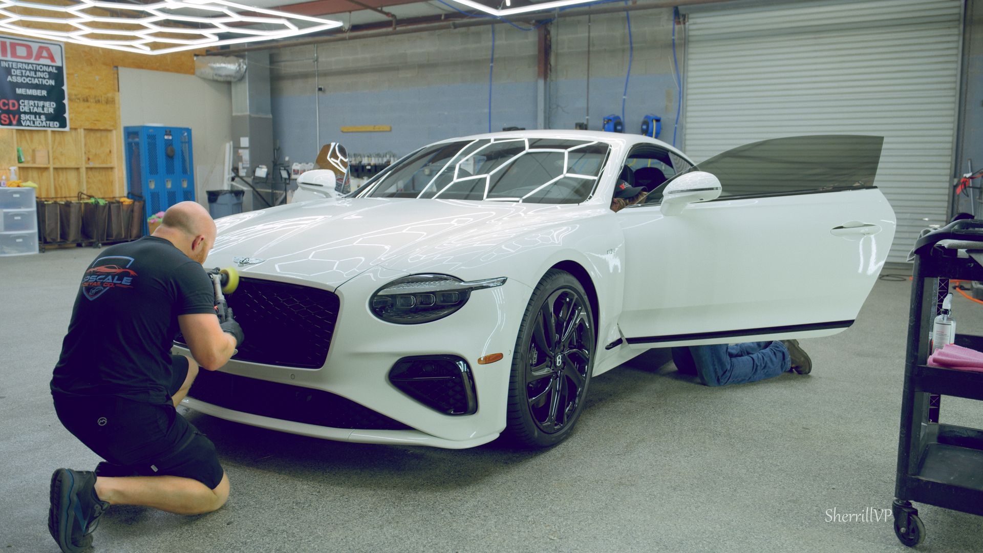 Man detailing a white Bentley in a garage, applying something to the front bumper. Another person works on the door.