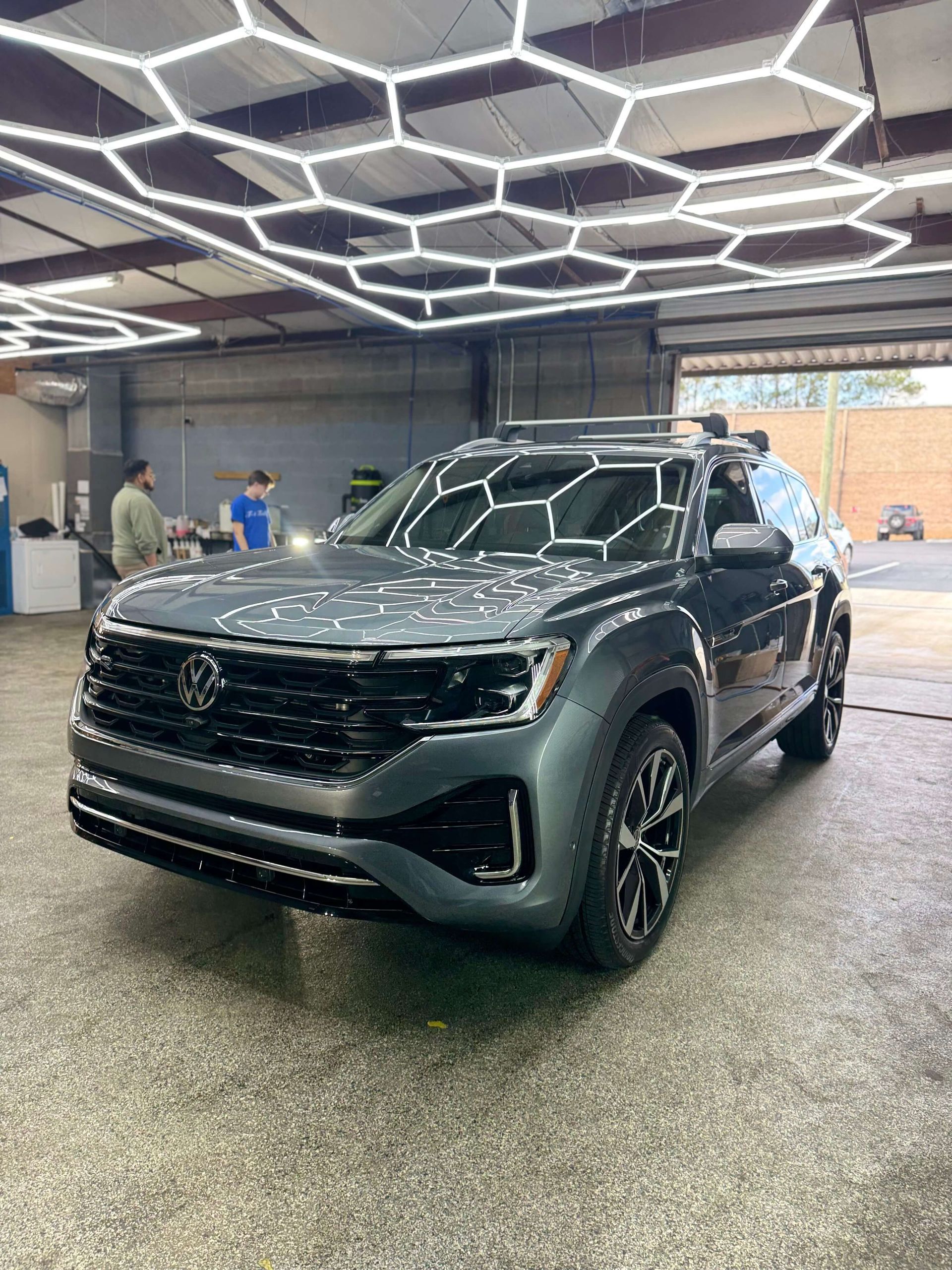 Gray Volkswagen Atlas SUV under hexagonal lights in a car detailing bay. Two people are in the background.