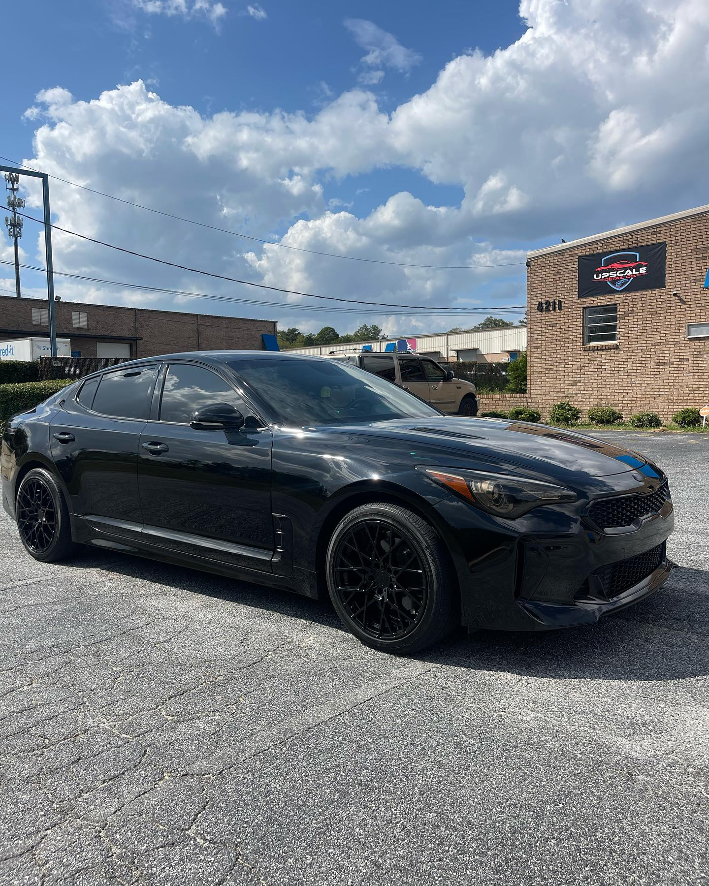 Black Kia Stinger parked on gravel, in front of a building with a blue sign, under a cloudy sky.