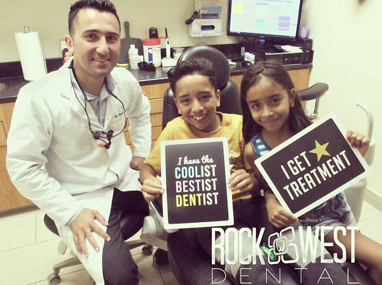 Dentist with two smiling children in a dental office. They hold signs that say "coolest dentist" and "I get treatment".