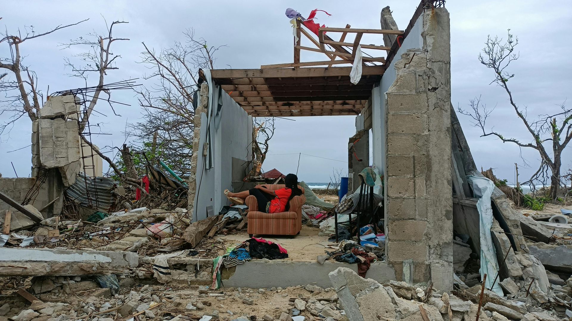 A woman is sitting on a couch in the ruins of a house.