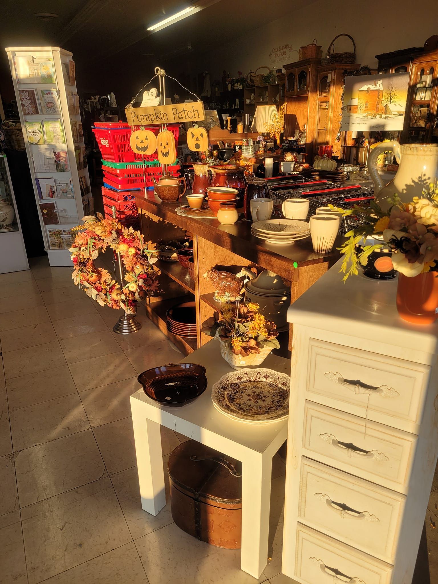 Inside a store, autumn-themed items displayed on tables and shelves. A white cabinet and small table are in the foreground.