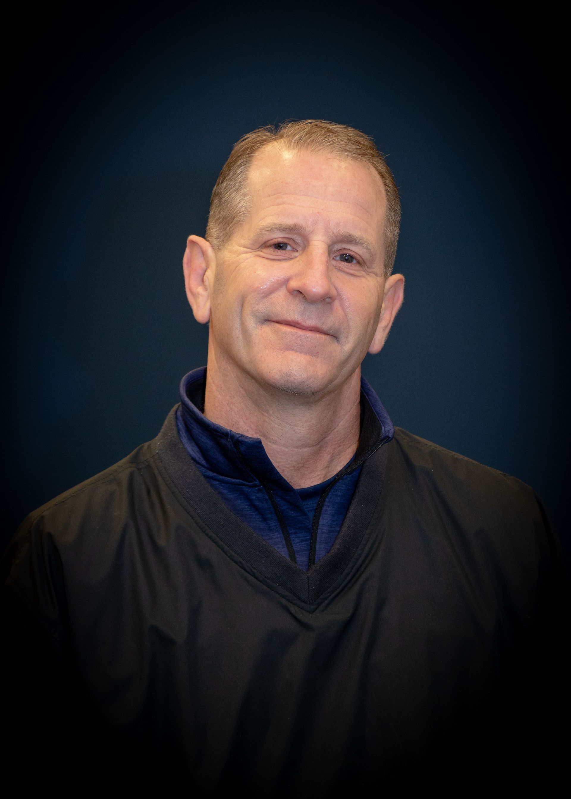 Man in black athletic top with blue collar, smiling in front of a dark blue background.