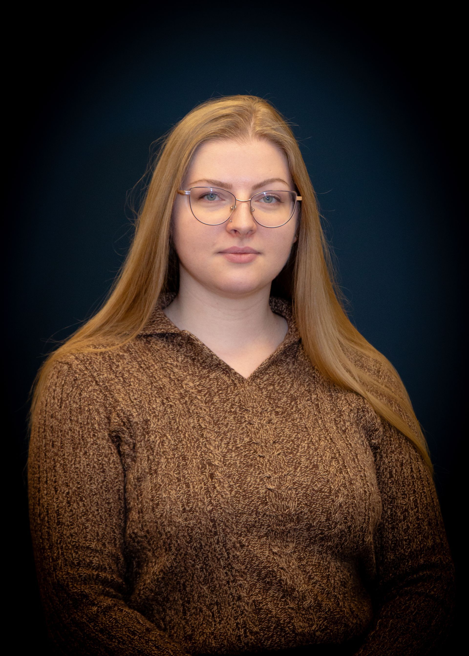Woman with long blonde hair and glasses, wearing a brown sweater, looking at the camera.