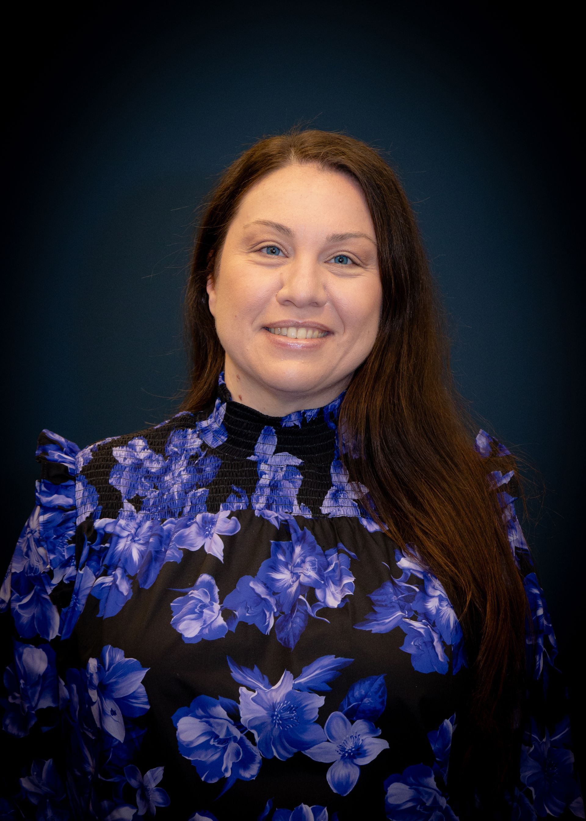 Woman with long brown hair, wearing a black floral print blouse, smiles at the camera against a dark blue background.