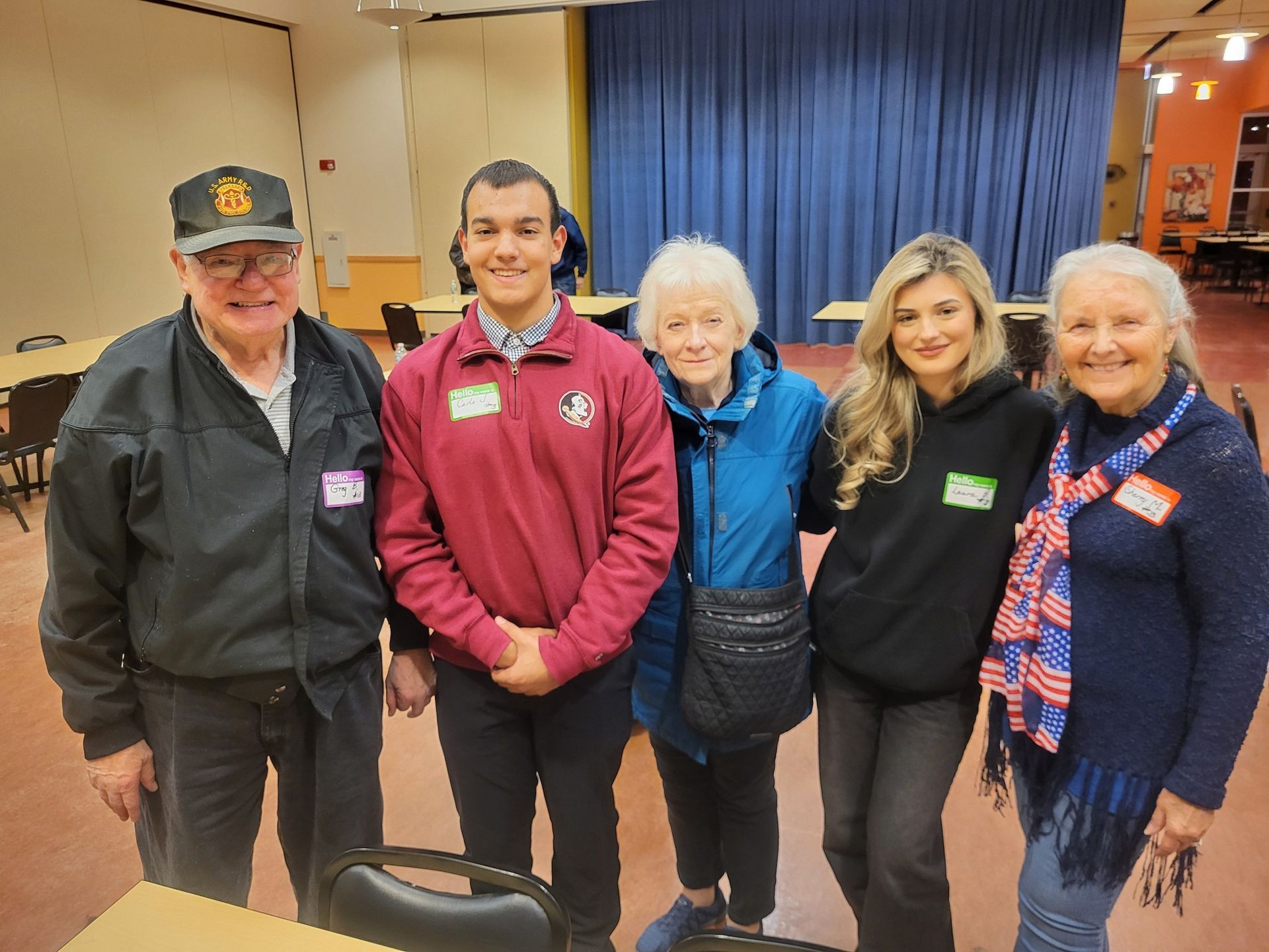 Five people smiling and posing indoors; one man wears a cap, another a burgundy shirt.