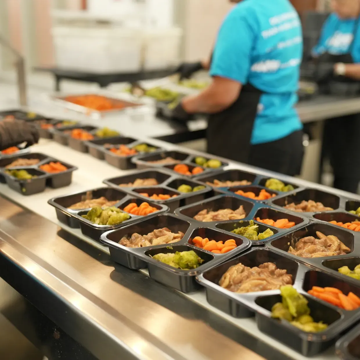 Prepared meal trays on a conveyor belt, with workers in blue shirts filling them with food.