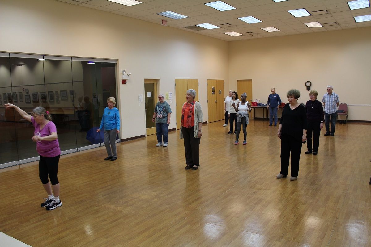People in a dance class, stretching and moving in a large room with wood floors.