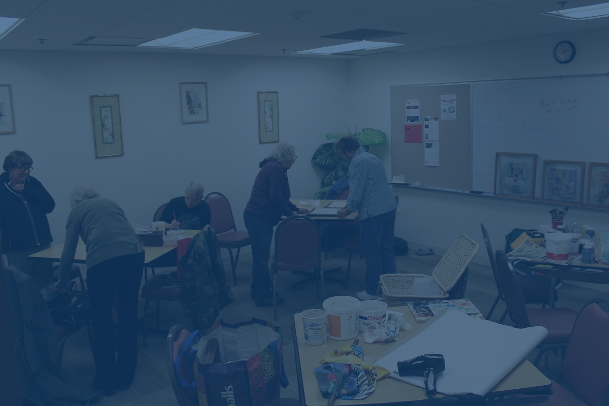 People at tables in a brightly lit room work on art projects. Paint supplies and artwork are visible.
