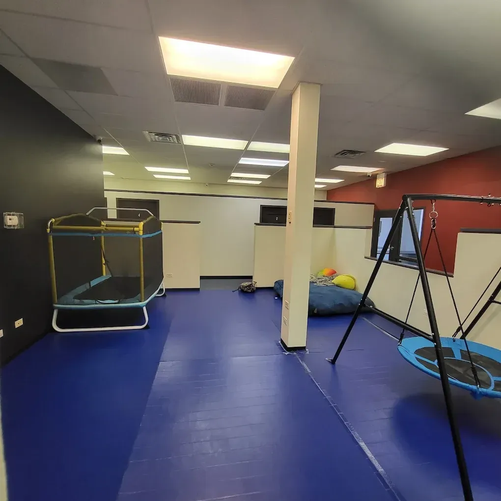 Indoor playroom with blue padded floor, trampoline, swing frame, and scattered bean bags under fluorescent lights